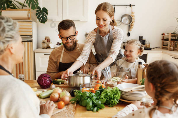 积极的年轻家庭主妇，带着小女儿，丈夫和祖母围坐在厨房桌子旁，准备美味的健康食品，并配上新鲜的配料，供全家在晚饭时享用