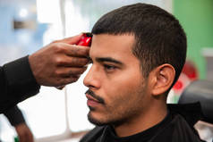 一名非洲裔美国人理发师在理发店里给一个留着山羊胡子和带着电动剃须刀的拉美裔拉美人理发.
