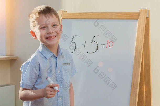 快乐，微笑的学龄前儿童站在一个白色的学校董事会上，手里拿着一个红色的记号和一个正确解题的数学例子。这个想法就是在家里教孩子们基本的学校知识