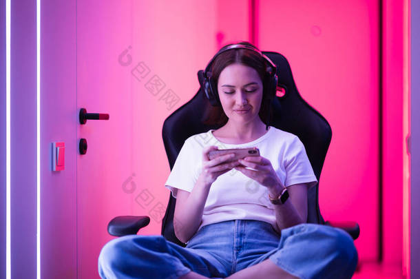网络游戏<strong>玩</strong>家坐在霓虹灯彩色房间的游戏椅子上，用智能<strong>手机玩手机</strong>游戏.