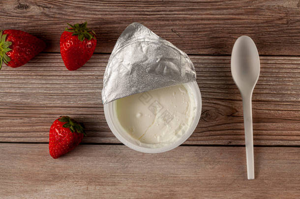 一个商店买了一杯纯低脂肪希腊酸奶，铝箔封皮脱落的平平铺图像。它的背景是木制的，有一个塑料勺子和新鲜的水果。饮食观念的概念