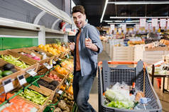 一个快乐的男人用手指指着购物车旁边的相机和超市里的食物 