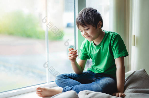 《快乐的孩子》的真实写照，笑脸坐在窗边，晨光下，活力充沛的男孩带着沉思的表情看着自己的手指，孩子在被监禁期间在家里自我封闭. 