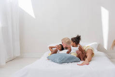 年轻的白人金发母亲玩耍，躺在白色的床上与非洲裔美国人的女儿的儿子。多民族家庭的快乐早晨。女人拥抱着,温柔地抚摸着孩子.简约的白色内部