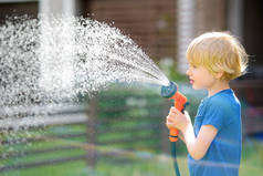 小男孩在草坪上浇水，在阳光充足的后院用花园软管与洒水器玩耍。学龄前儿童与喷水为乐.孩子在帮他的父母家庭抚养。教孩子工作吧