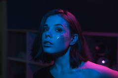 一个穿着化妆品的漂亮女人的画像，在室内蓝色和紫色的灯光下闪烁着光芒。晚上派对上的女人看着镜头.