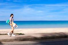 年轻女子带着冲浪滑板在热带海滩附近的小道上行走。小女孩长发，身穿长袖衬衫，牛仔裤和运动鞋，脚踏着冲浪滑板在海滩路上行走.