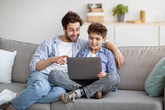 照顾爸爸教儿子一起使用笔记本电脑或上网，坐在客厅舒适的沙发上