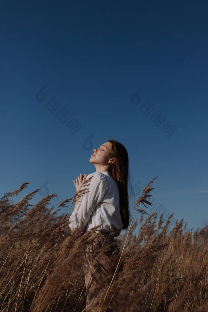 年轻的女人，穿着白衬衫，紧闭双眼，站在蓝天和太阳前干枯的草原上。都市风格和街头时尚。穿休闲装的女孩抬头仰望天空