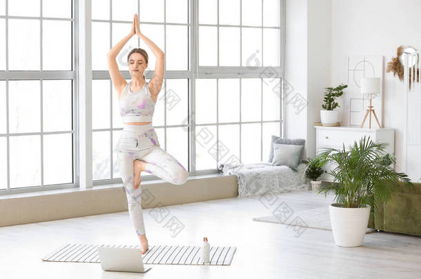 一个身材矮小的年轻女子，带着笔记本电脑，在家里练习瑜伽