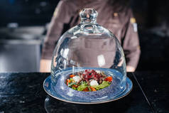 一名专业厨师在一个玻璃圆顶下精致的餐馆里，用新鲜准备的西红柿和小牛肉蔬菜沙拉配酱汁.