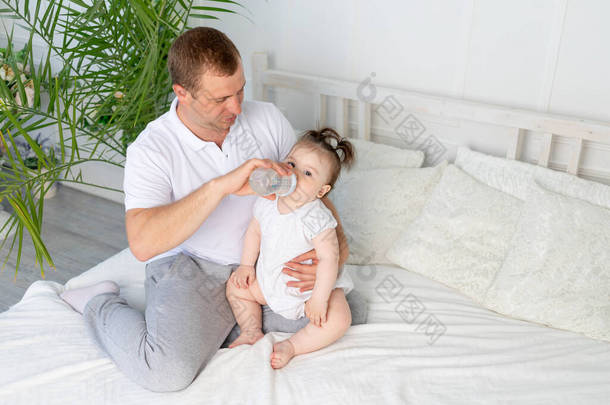一位父亲在家里的白色床上用瓶子喂养他的小女儿，这是婴儿<strong>食品</strong>和<strong>儿童</strong>保育的概念