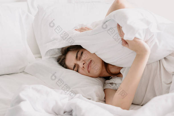 睡眠质量差的概念。年轻的愤怒而疲惫的女人用枕头捂住耳朵，淹没了噪音，无视警报，试图在明亮的卧室里醒来