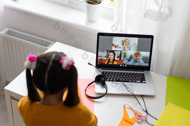 可爱的小女生在线聊天Skype笔记本电脑在线课上的视频电话课桌使用耳机向同学致意挥手隔离研究客厅室内.