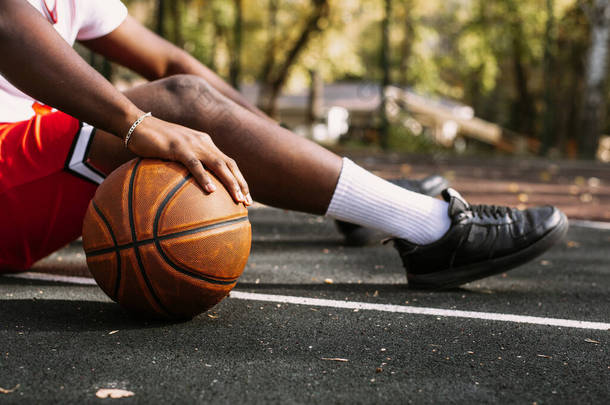 一个手握篮球、坐在篮球场上的黑人年轻人的特写。运动期间休息一下