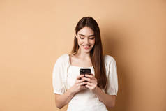 年轻女子在手机上发短信，微笑着阅读智能手机屏幕，站在米色背景上