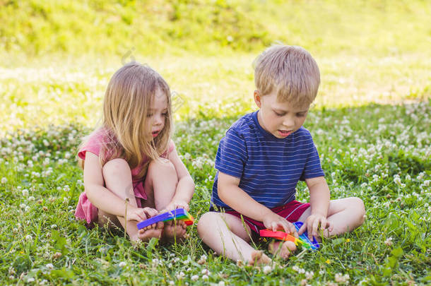 金发男孩和女孩坐在绿草草坪上玩硅胶玩具减压弹出。弹出它感官玩具。压力缓解。彩色抗压硅酮传感器玩具
