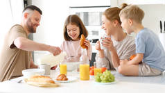 年轻快乐的家庭，家里有两个可爱的小孩在厨房里吃着健康的早餐，笑着的父亲把牛奶倒在玻璃瓶里，而孩子们的女儿和儿子在等着吃早餐