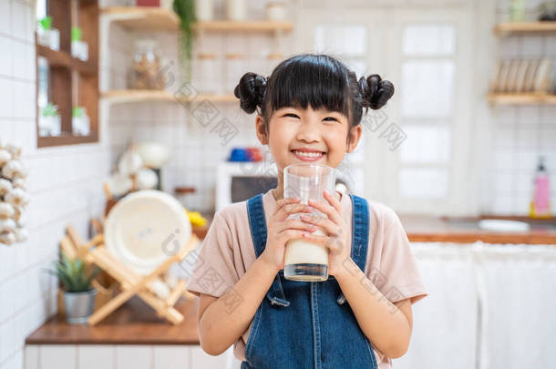 在家里厨房里拿着一杯<strong>牛奶</strong>的亚洲可爱小孩的画像。学龄前小女孩或女儿笑着呆在家里，开心地<strong>喝牛奶</strong>，然后看着相机.