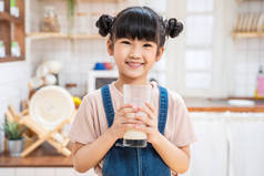 在家里厨房里拿着一杯牛奶的亚洲小孩的画像。幼稚园可爱的小女孩或女儿笑着呆在家里，开心地喝牛奶，然后看着相机.