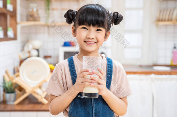 在家里厨房里拿着一杯牛奶的亚洲小孩的<strong>画像</strong>。幼稚园可爱的小女孩或女儿笑着呆在家里，开心地喝牛奶，然后看着相机.