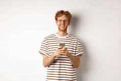 技术和电子商务概念。戴眼镜的红头发男人用手机笑着。拿着智能手机的年轻人高兴地盯着摄像机，白色背景