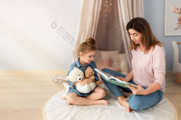 一位中年妇女正在儿童房给女儿读书。那姑娘<strong>手里</strong>拿着一只红白相间的熊.
