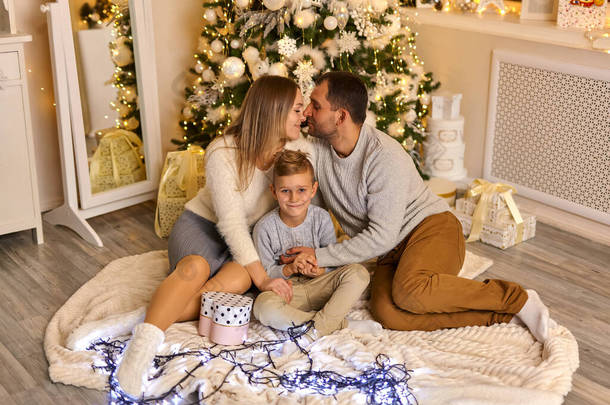 快乐的父母和可爱的小儿子坐在地板上，在圣诞树的背景上装饰着新年灯室。家庭、假日概念