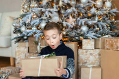 背景树上被包裹着的圣诞礼物让学龄前的男孩感到兴奋。圣诞快乐和节日快乐的概念。孩子在家里打开礼物。孩子很有兴趣也很惊讶
