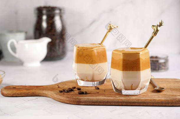 两杯带有时髦韩国饮料达拉那<strong>咖啡</strong>拿铁的玻璃杯- -<strong>速溶咖啡</strong>，砂糖，热水和牛奶在木板上，白色表面