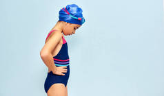 一个戴着护目镜，身穿泳衣，头戴一顶泳帽，在游泳前朝下看的可爱小女孩的横向侧视图，孤立的蓝色摄影棚背景.