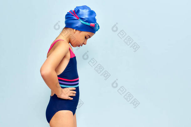 一个戴着护目镜，身<strong>穿泳衣</strong>，头戴一顶泳帽，在游泳前朝下看的可爱<strong>小女孩</strong>的横向侧视图，孤立的蓝色摄影棚背景.
