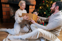 幸福的年轻女子坐在壁炉旁和圣诞树旁送丈夫和儿子圣诞礼物的画像.
