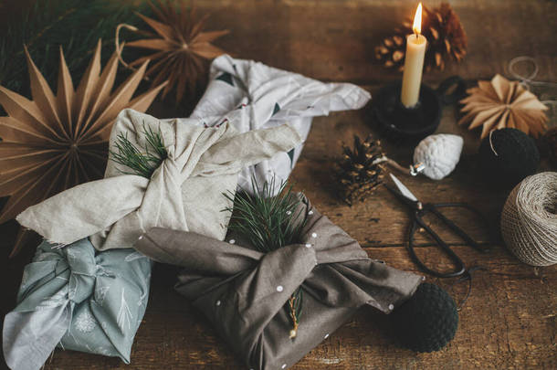 圣诞礼物用面料包裹在乡村木桌上，上面有剪刀、纸星、蜡烛、装饰品。大气喜怒无常的图像，<strong>北欧风</strong>格。圣诞快乐！Furoshiki包，零浪费假期