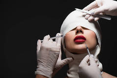 young woman in elastic bandage near plastic surgeons with surgical knife and syringes isolated on gr