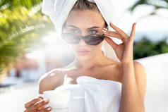 年轻迷人的高加索女人洗澡或洗澡后在阳台上享受户外咖啡。穿着白色毛巾，头戴时髦太阳镜，头戴咖啡壶或茶的女孩。奢华的生活方式放松点旅游业.