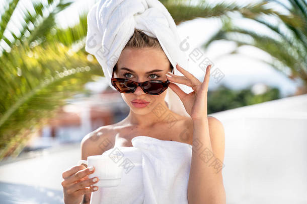 年轻迷人的高加索女人洗澡或洗澡后在阳台上享受户外咖啡。穿着白色毛巾，<strong>头戴</strong>时髦太阳镜，<strong>头戴</strong>咖啡壶或茶的女孩。奢华的生活方式放松点旅游业.