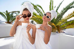 两个穿着白色毛巾的迷人的高加索女人在宾馆里享受温泉日，在阳光明媚的夏日放松，在阳台上喝咖啡。背景上的棕榈奢华的生活方式友谊。女朋友们。旅游业