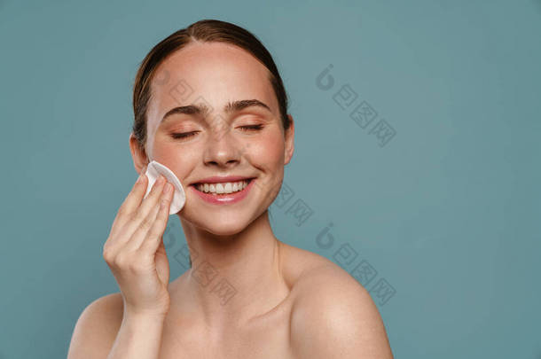 Happy young woman cleancing her face with cotton pad over blue background