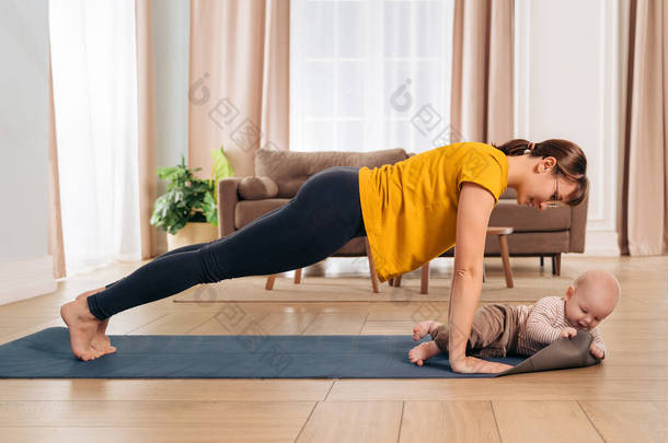 Beautiful yogi <strong>mom</strong> smiling at her baby while doing push up on exercising mat at home. Happy <strong>mom</strong> work