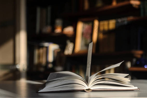 There is an <strong>open</strong> book on the table.  The background is bookshelves illuminated by the sun.