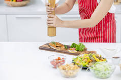 在厨房里，红围裙女士的手将调味品撒在用不同配料和装饰的鱼片盘上。在他们家里做饭快乐的概念.
