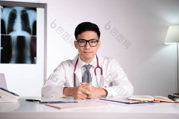 在医院的诊疗室里，年轻、友善、礼貌的亚洲医生、骨科医生，戴着听诊器，手持笔记本电脑，直视身后的X光图像。医疗设施中的亚洲合格医生，注意和注意