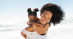 快乐的黑人年轻女人一边笑一边在沙滩上给女儿搭便车。可爱的非洲小女孩，姐姐在海滨户外玩耍。一个漂亮无忧无虑的妈妈紧紧地抱着她那可笑的大腿骨