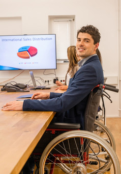 包容肖像男人。轮椅包容肖像男人。轮椅参与会议与的同事们办公室环境与屏幕的背景显示国际销售数据