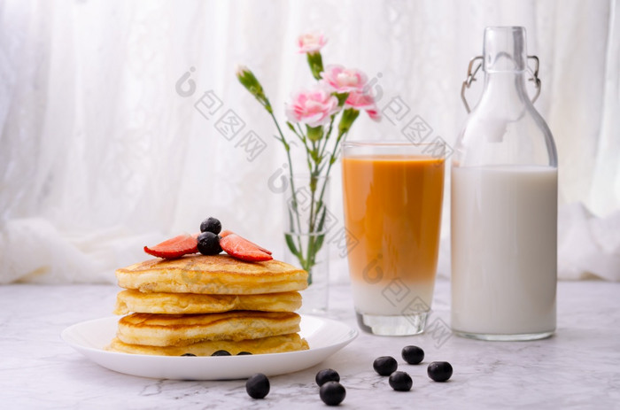 堆栈煎饼与新鲜的草莓和蓝莓白色板瓶牛奶玻璃牛奶茶和花瓶粉红色的花大理石表格健康的早....早餐