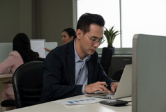 年轻的亚洲商人眼镜工作移动PC他的桌子上开放空间办公室与的同事们的背景