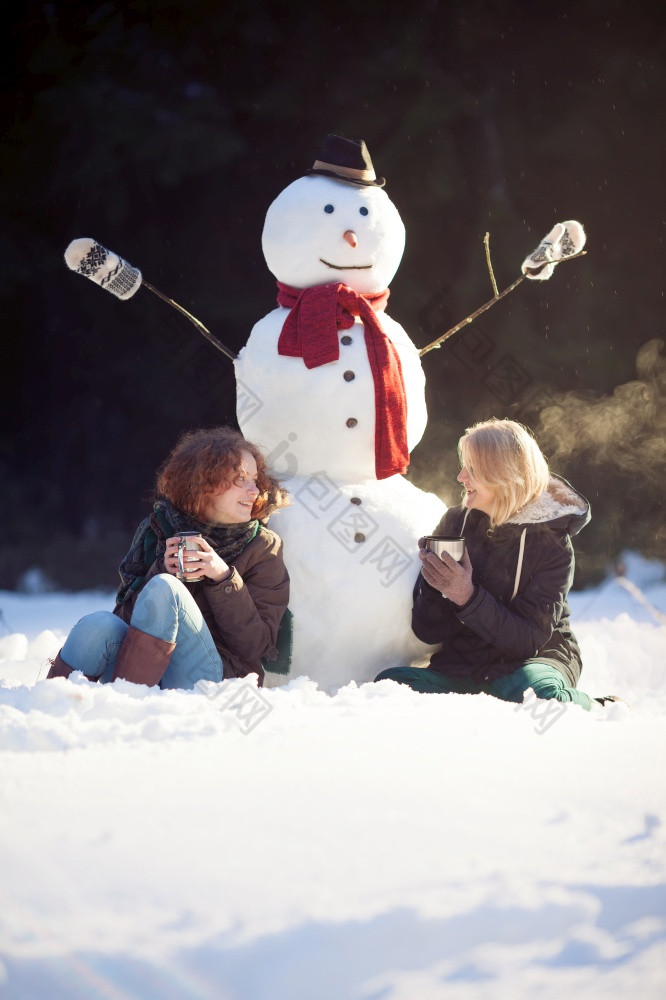 两个年轻的女人坐着附近雪人和喝热茶阳光明媚的冬天一天焦点女孩