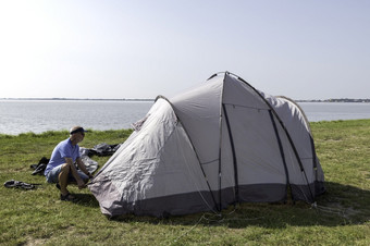 成熟的男人。设置帐篷和把下来帐篷挂钩场附近的水美丽的空间附近的湖男人。设置帐篷的水线