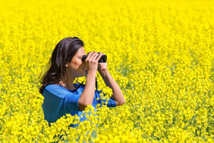女人看通过双筒望远镜盛开的油菜籽场年轻的女人看通过双筒望远镜开花黄色的油菜籽场